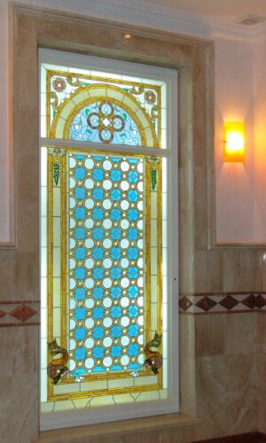Roman Design Stained Glass Window with blue and gold geometric patterns set in beige marble, beside a lit wall sconce.