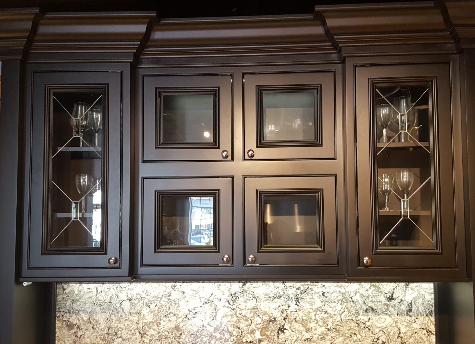 Astoria dark wood cabinets with glass panel doors display glassware above a granite countertop backsplash.
