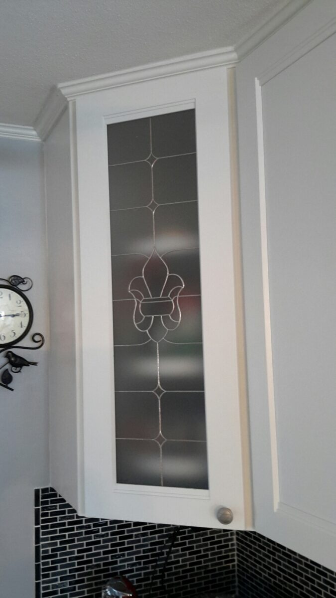 White kitchen cabinet with a Fleur De Lis glass insert, featuring geometric and fleur-de-lis center designs.