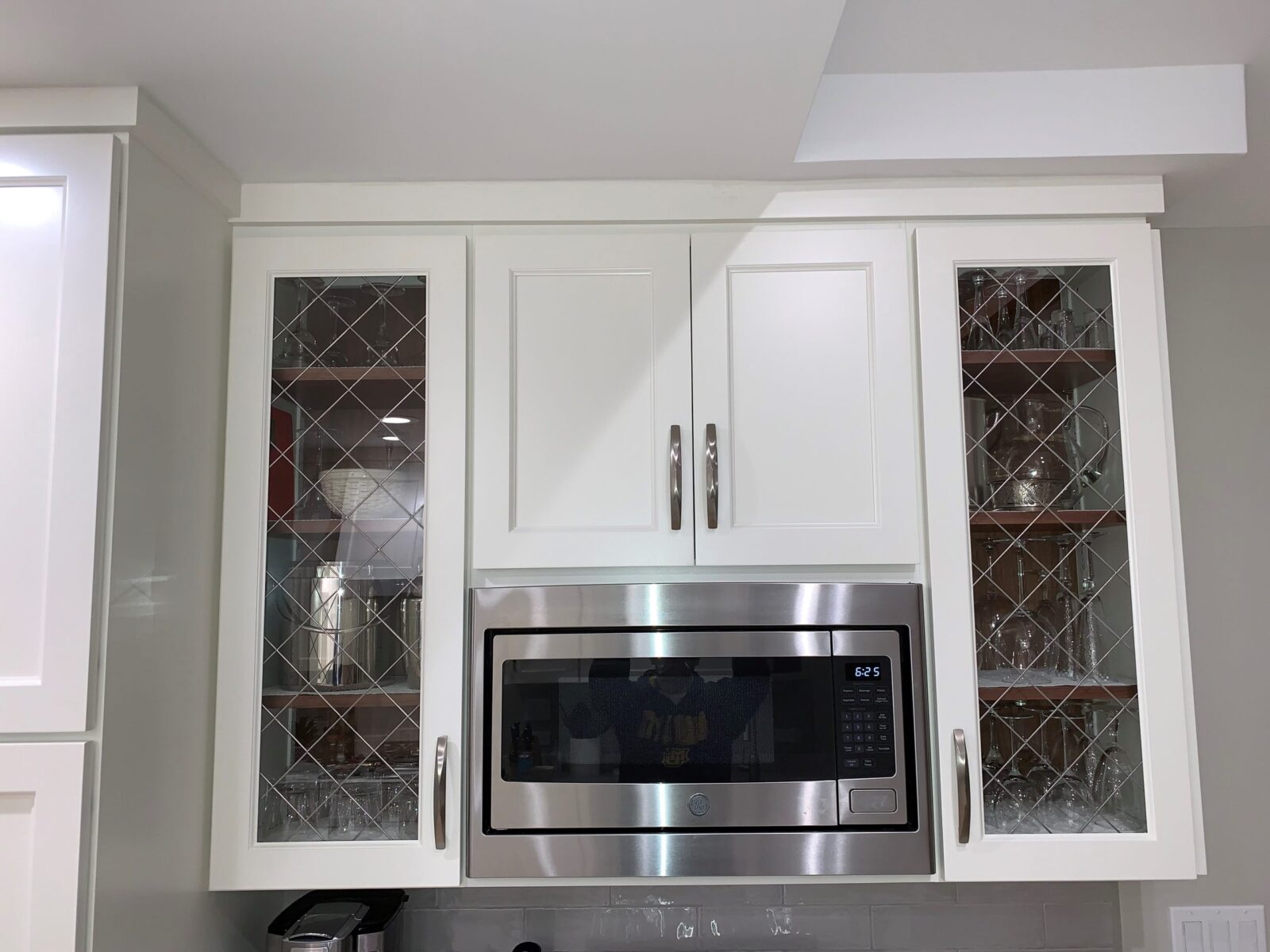 White kitchen cabinets with Apollo glass doors flank a stainless microwave above a tiled backsplash.