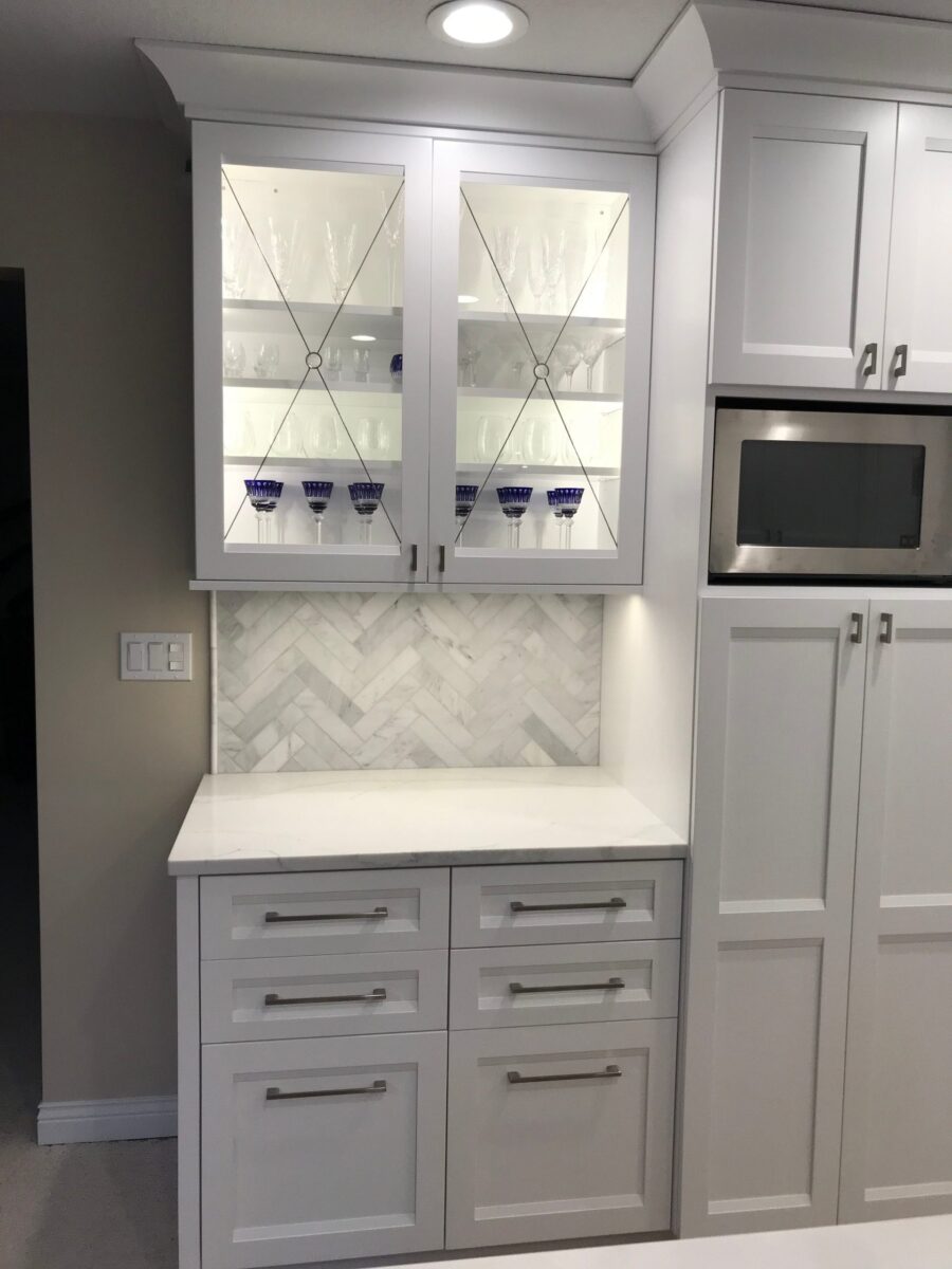 Chester cabinetry with glass inserts displays glassware above a marble herringbone backsplash.