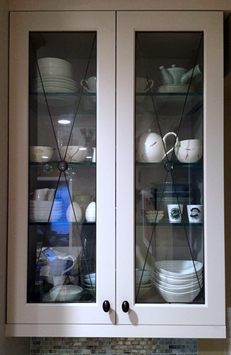 Chester cabinet with glass inserts displays organized dishes, bowls, cups, and teapots on three shelves.