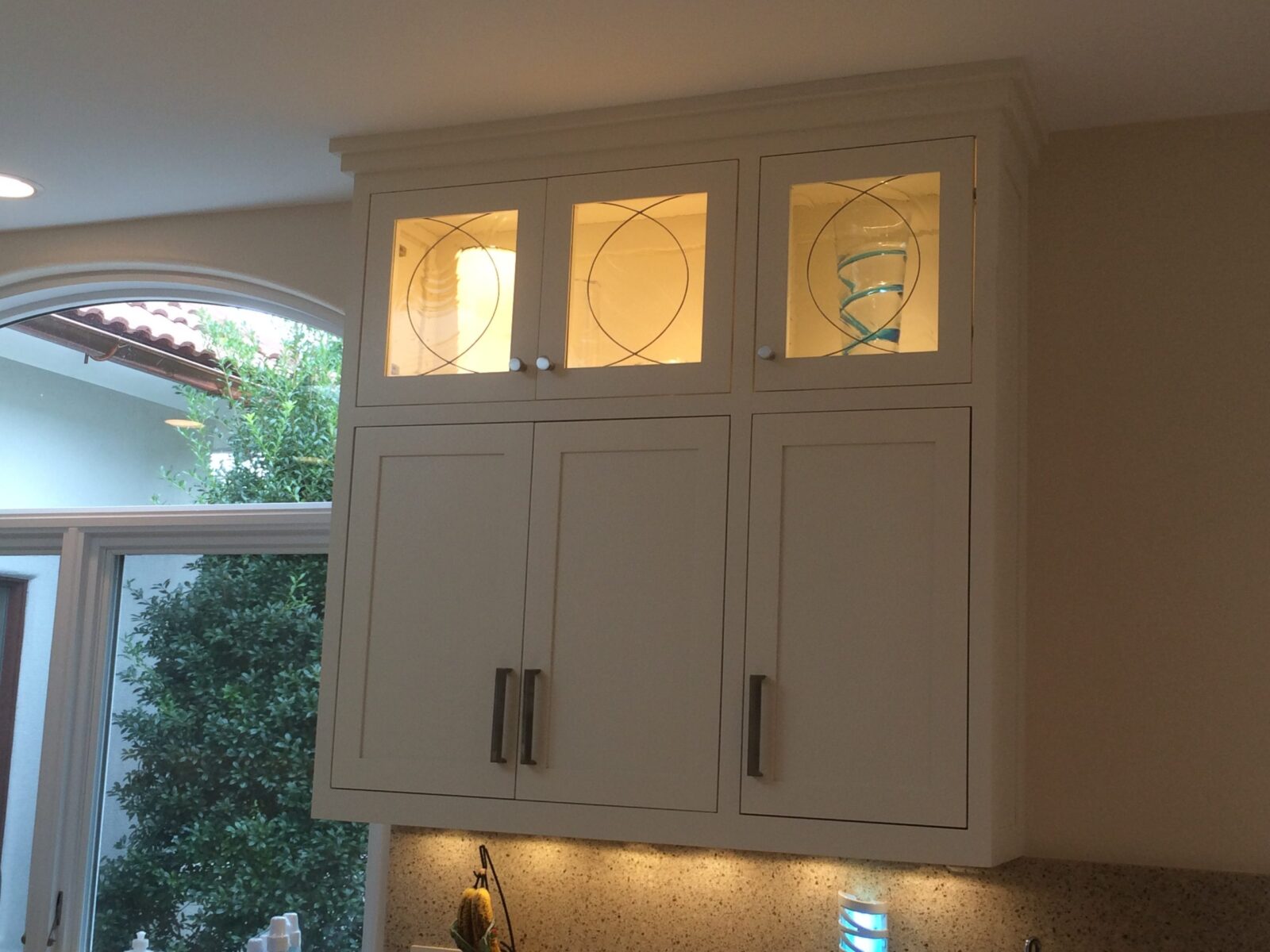 White kitchen cabinet with Da Vinci glass inserts, glass doors, and lit shelves for displaying decor.