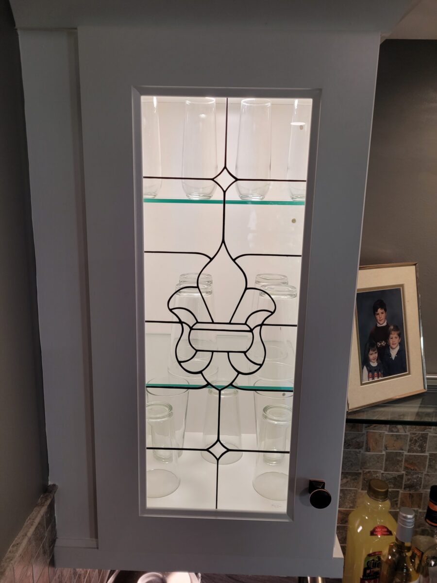 White cabinet with a Fleur De Lis glass insert door displaying empty jars and glasses inside.
