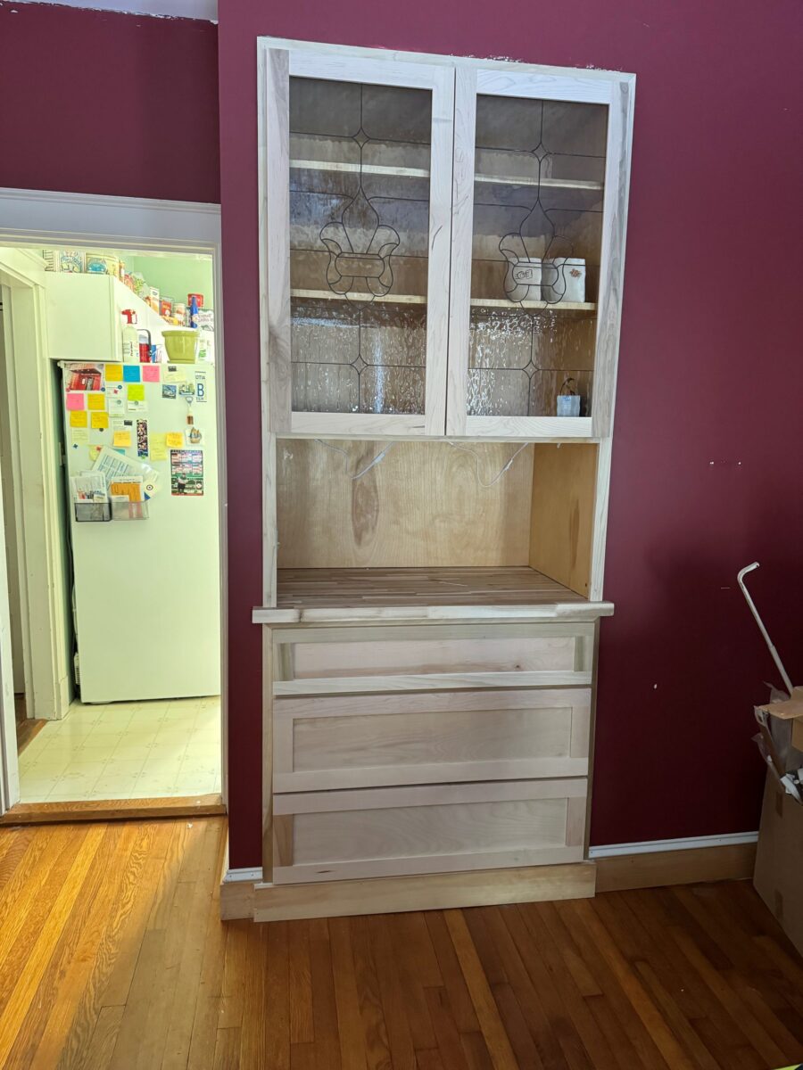 Fleur De Lis unfinished cabinet with glass doors and drawers stands against a red wall near the kitchen.