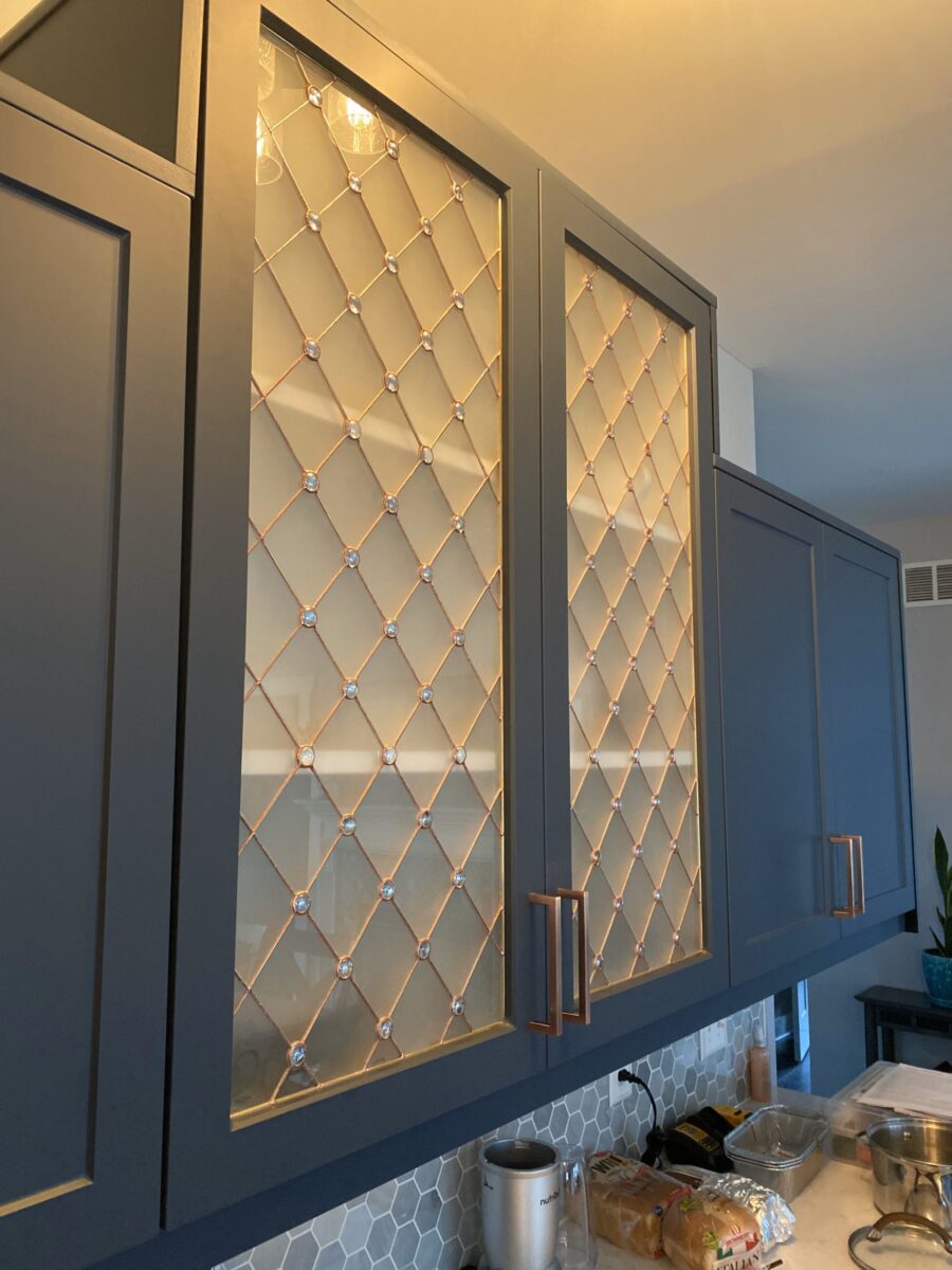 Savannah blue cabinets with diamond art glass inserts and copper handles above a counter full of food.