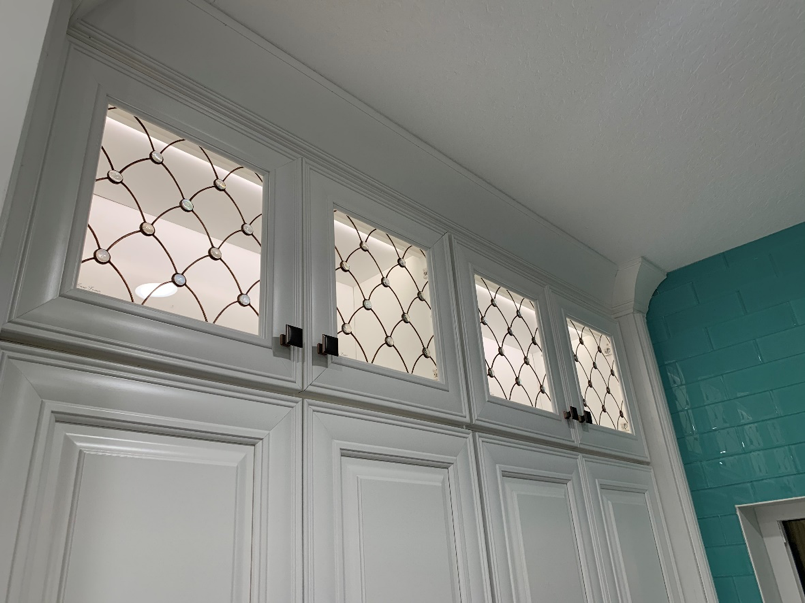 Venezzia white cabinets feature art glass and metal grilles, beautifully set against a turquoise tiled wall.