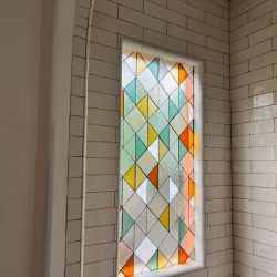 Stained glass window with orange, yellow, green, and clear diamond shapes set in a white tiled wall.