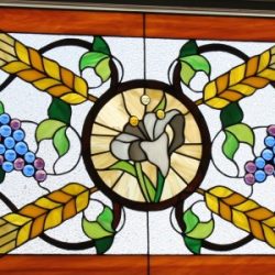 Stained glass window with a white flower, wheat stalks, and grape clusters in a symmetrical design.