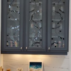 Gray glass-front cabinets with geometric patterns above a marble countertop displaying decor, fruit, and a framed picture.