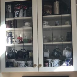 White kitchen cabinet with glass doors displaying assorted mugs, cups, and glasses arranged on shelves inside.