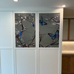 Stained glass panels with birds and flowers set in white cabinetry, under recessed ceiling lights in a kitchen area.