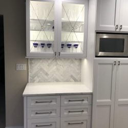 White kitchen cabinets with glass doors display glassware above drawers and a marble tile backsplash.