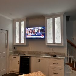 A kitchen with white cabinets, a wall-mounted TV displaying news, and a marble countertop in the foreground.