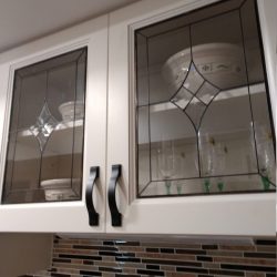 White kitchen cabinets with glass doors display bowls and glasses above a tiled backsplash in brown and beige tones.