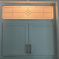 White cabinet with two doors and silver handles, topped by a rectangular stained glass window with geometric patterns.