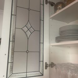 Open cabinet with a glass-paneled door, stacked plates, glasses, a wine glass, and a small decorative item inside.