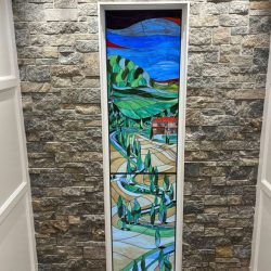 Tall stained glass window showing a landscape with hills, winding roads, trees, and a red-roofed house against a stone wall.