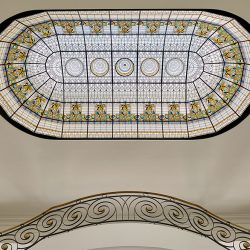 Ornate stained glass ceiling above a curved staircase with an intricate metal railing in a bright interior.
