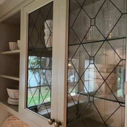 Glass-front cabinet with lattice design displays plates, bowls, and glasses on shelves; window reflection is visible.