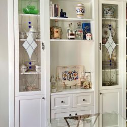 White cabinet with glass doors displays glassware, ceramics, and bottles; shelves hold decorative and bar items.
