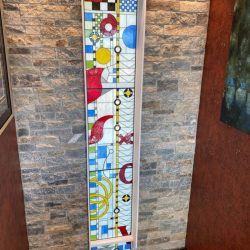 Tall stained glass window featuring geometric shapes, bold colors, and abstract patterns set in a stone wall.