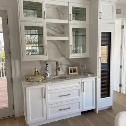 White kitchenette with glass cabinets, a small sink, gold faucet, and a built-in wine fridge on light wood flooring.