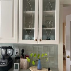 White kitchen cabinets with glass doors, a counter with a blender, utensils, a plant in a vase, and a small bottle.