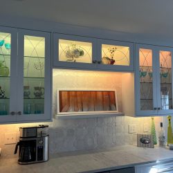 Glass-front cabinets with lighted shelves display glassware above a countertop with bottles and a coffee maker.