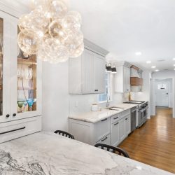 Modern kitchen with white cabinetry, marble countertops, glass display cabinet, and a bubble chandelier over a large island.