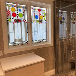 Two stained glass windows with geometric patterns next to a light wood vanity and glass shower in a modern bathroom.