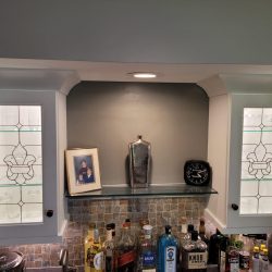 Glass-front cabinets and a shelf with a framed photo, silver bottle, and clock above a counter with various liquor bottles.