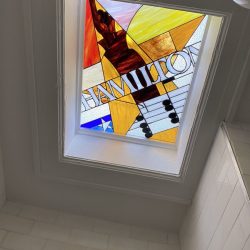 Stained glass skylight with abstract design, musical notes, and the word “HAMILTON” visible in bold letters.