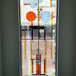 Stained glass window with geometric shapes and colored glass in a modern, abstract style, framed by white casing.