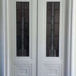 Closed double doors with geometric dark glass panels and white molded frames, set against a neutral wall.