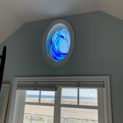 Round stained glass window with blue wave pattern above a door overlooking a sandy outdoor area and beach.