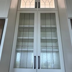 Tall white kitchen cabinet with glass doors, black handles, and interior shelves, set against a tiled backsplash.