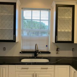 Modern kitchen with glass-front cabinets, a black countertop, and a sink positioned below a window with blinds.