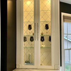 White cabinet with glass doors, displaying black goblets and decorative items, set against a dark wall near a window.