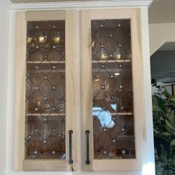 Wooden cabinet with glass-paneled doors, displaying glassware on shelves inside. Plants visible on the right side.