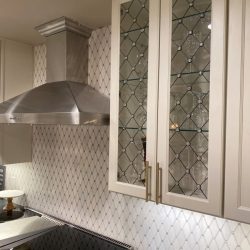 Glass-front kitchen cabinets with a geometric pattern beside a stainless steel range hood and tiled backsplash.