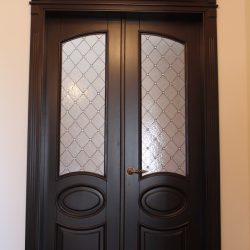 Dark wooden double doors with decorative frosted glass panels and brass handles, set in a neutral-colored wall.