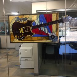Stained glass art of an electric guitar with mountains, hung in an office with glass walls and visible desks.
