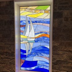 Stained glass window depicting a white sailboat on blue water with colorful fields and sky in the background.