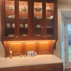 Wooden kitchen cabinet with glass doors displaying dishes, marble countertop, and small decor items underneath.