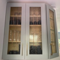 Gray kitchen cabinet with textured glass doors, displaying dark dishes inside, positioned in a sunlit corner.