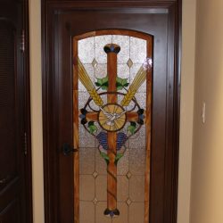 Wooden door with a stained glass panel featuring a cross, wings, and grapevine design, set in a beige wall.