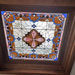 Stained glass ceiling panel with geometric and floral patterns in blue, brown, and pink, framed by dark wood.