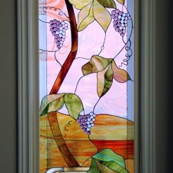 Stained glass window depicting a grapevine with green leaves and purple grapes in a white vase against a pastel background.