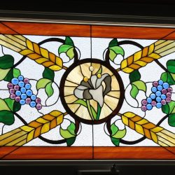 Stained glass window with wheat, grapes, green leaves, and a central white flower on a geometric background.